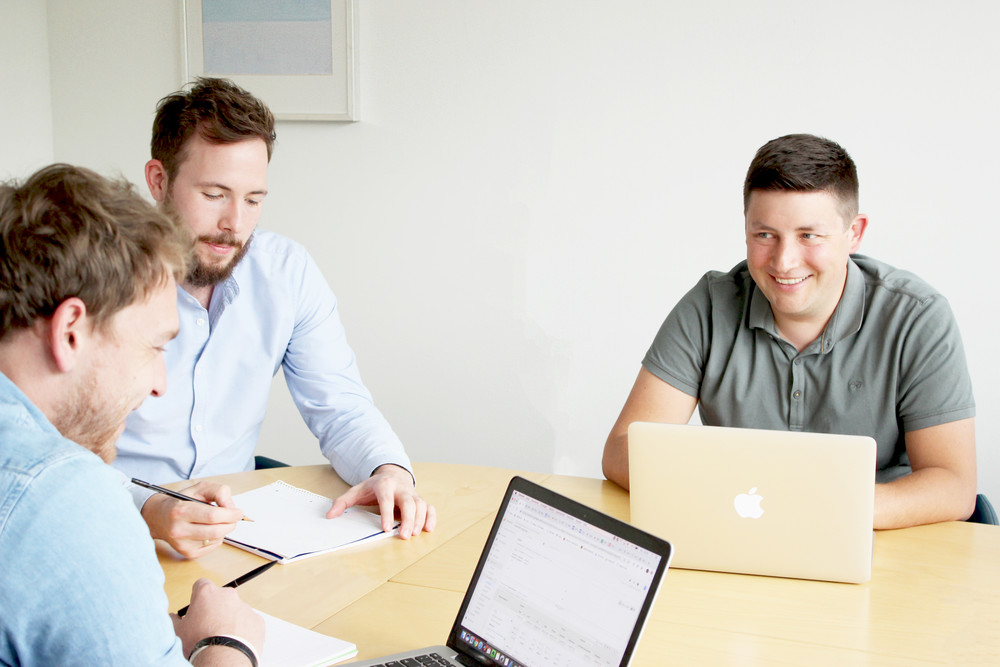 Drei tms Kollegen sitzen um einen Konferenztisch. Auf dem Tisch stehen zwei geöffnete Laptops sowie zwei Notizblöcke.